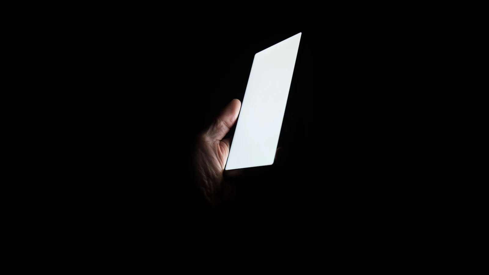 A hand holding a smartphone in the dark, its blank bright screen the only light in the frame